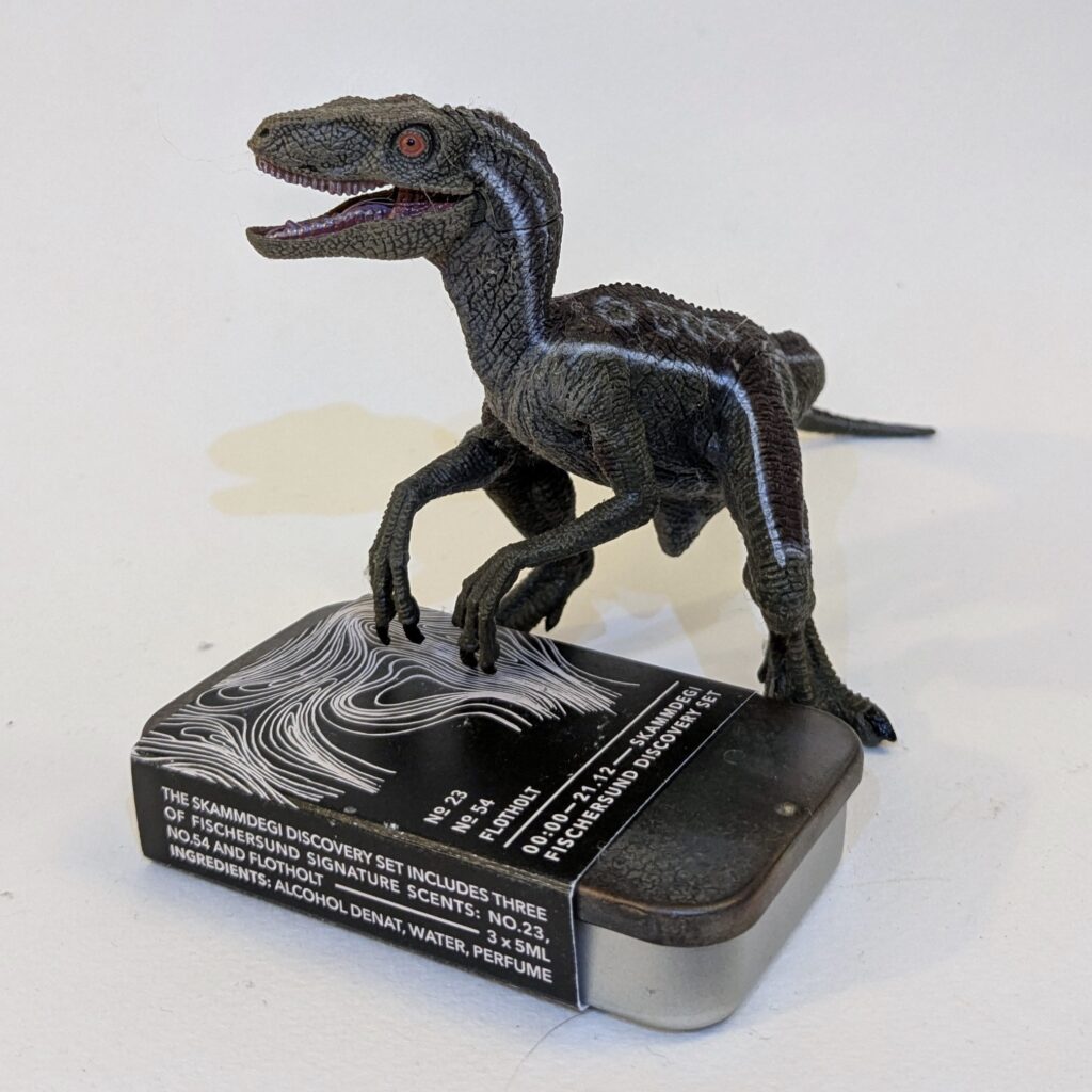 A small grey plastic velociraptor toy, jaws open, standing with its front claws on a metal tin of Fischersund perfume samples