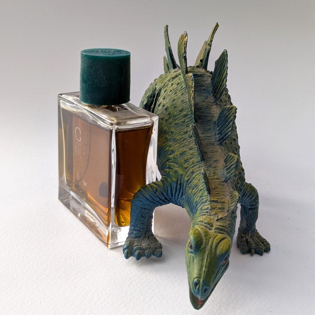 A green and blue plastic stegosaurus standing next to a flat square bottle of Bogue perfume